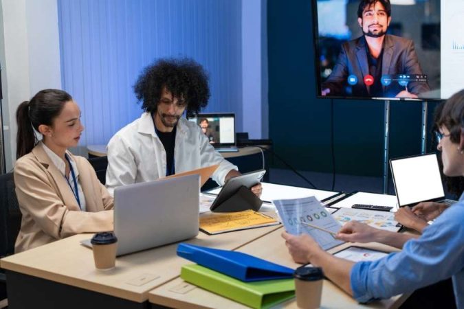 Une équipe de bureau réunie autour d'une table, analysant des données et discutant lors d'une visioconférence avec un collègue sur un grand écran.
