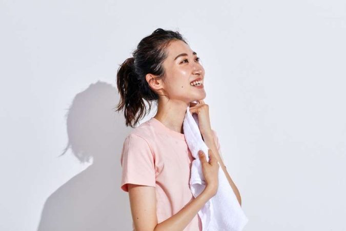 Une jeune femme souriante tient une serviette blanche contre son cou dans une ambiance lumineuse.