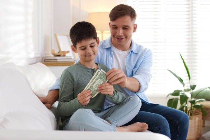 Un père souriant donne de l'argent de poche à son jeune garçon assis sur un canapé, illustrant le versement d'argent de poche à son enfant.