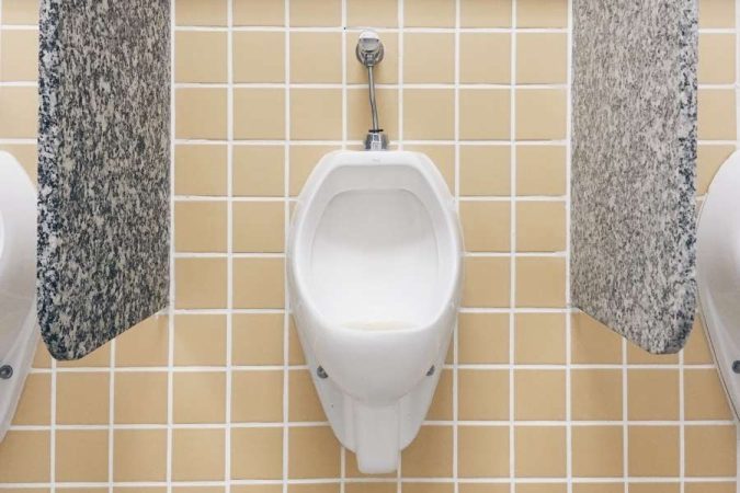 Gros plan sur un robinet de chasse d'eau en métal chromé actionné par un bouton poussoir, installé au-dessus d'un urinoir mural en céramique blanche dans des toilettes publiques carrelées de jaune.