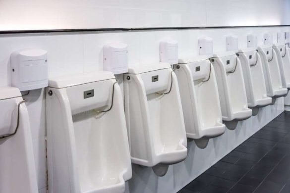 Rangée de plusieurs urinoirs muraux modernes en céramique blanche avec des distributeurs de désinfectant fixés au mur au-dessus, dans des toilettes publiques propres avec carrelage blanc et sol sombre.